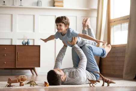 Joyful young man father lying on carpet floor, lifting excited happy little child son at home. Full length carefree two generations family having fun, practicing acroyoga in pair in living room.